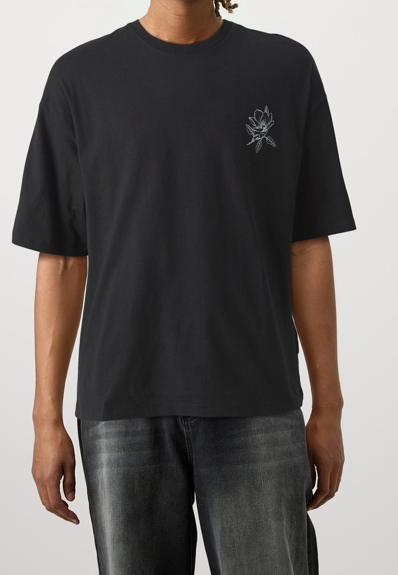 Svart bomullströja med korta ärmar, prydd med en liten vit blommig broderi på vänster bröst. Enkel rund halsringning och tyg som sitter löst.