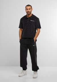 Man standing with hands in pockets wearing black textured polo shirt and matching pants with white sneakers on plain background.
