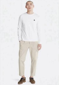 Weißes Langarm-T-Shirt mit kleinem schwarzem Logo, kombiniert mit beigen, zulaufenden Hosen und braunen Schnürschuhen. Einfaches, lässiges Outfit.