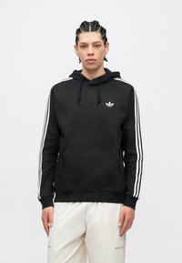 Jaunas žmogus su pintinėmis kasomis, dėvintis juodą Adidas džemperį su baltomis juostelėmis ir baltas kelnes, stovintis priešais paprastą foną.