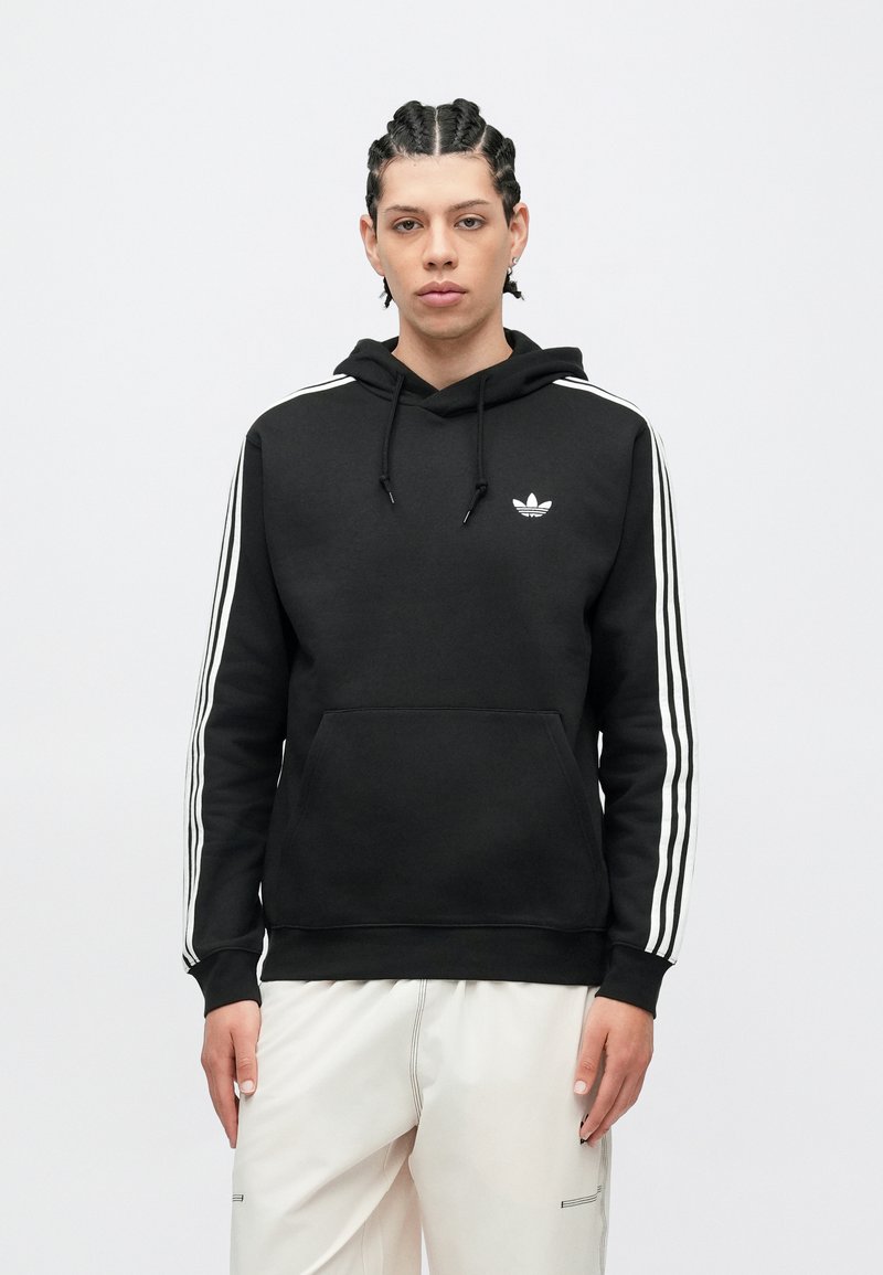 Giovane con capelli intrecciati che indossa una felpa nera Adidas con strisce bianche e pantaloni bianchi, in piedi davanti a uno sfondo semplice.