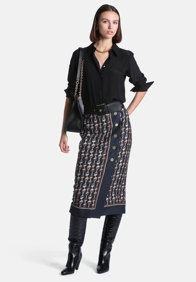 Femme portant une chemise noire boutonnée, une jupe midi à motifs avec des boutons, des bottes hautes noires, et portant un sac à main noir sur une épaule.