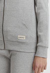 Siva zip-up dukserica napravljena od teksturirane tkanine, koja ima bočne džepove i oznaku branda. Rukavi su uski, a dno je u istom sivoj boji.