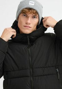 Jachetă cu puff neagră, cu guler înalt, fermoar frontal și buzunare laterale. Modelul poartă o căciulă tricotată de culoare gri deschis, cu un etichetă.