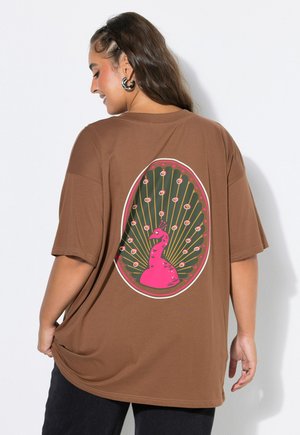 Donna che indossa una t-shirt marrone oversize con una grafica colorata rosa e verde di un pavone sulla schiena, in piedi contro un muro bianco.