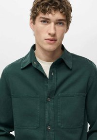 Camicia button-up di un verde scuro, realizzata in un tessuto resistente; presenta tasche sul petto e un colletto classico. Texture morbida, design pulito.