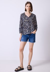 Bluson mit langen Ärmeln und floralem Muster, mit Rüschenausschnitt, kombiniert mit Jeansshorts mit umgeschlagenen Säumen. Schwarze Sandalen mit silbernen Hardware-Akzenten.
