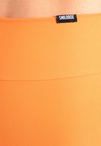 Oranssi joustava kangas, jossa sileä pinta. Vyötäröllä musta merkki, jossa lukee "SMILODOX". Yksinkertainen, istuva muotoilu.