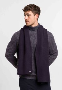 Navy knitted scarf made of thick fabric, featuring a textured pattern. Worn over a gray chunky turtleneck sweater, showing a smooth finish.