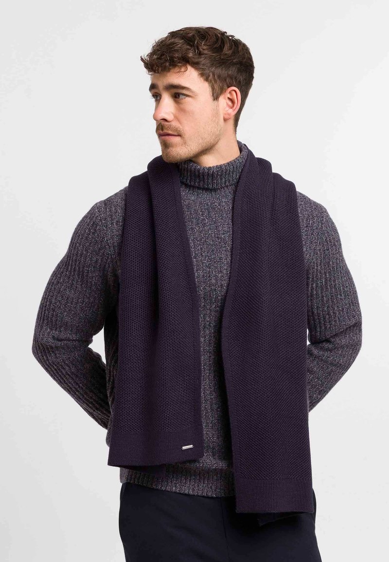 Navy knitted scarf made of thick fabric, featuring a textured pattern. Worn over a gray chunky turtleneck sweater, showing a smooth finish.