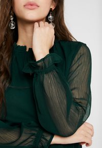 Blusa verde scuro con maniche lunghe trasparenti e testurizzate. Presenta un colletto e polsi arricciati, completati da orecchini trasparenti.
