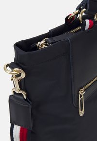 Borsa nera in materiale sintetico con dettagli dorati, finitura testurizzata, una tasca con zip e una tracolla striata con accenti rossi, bianchi e blu.