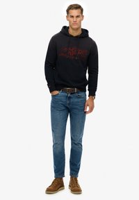 Marineblå hættetrøje med et rødt grafisk logo, parret med lyseblå denimjeans og brune snørebåndsstøvler, med et bælte og ur.