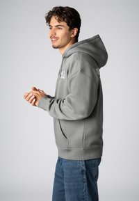 Gray hooded sweatshirt with a soft texture, kangaroo pocket, and ribbed cuffs. Printed logo detail on the front and back. Paired with blue jeans.