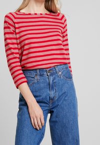 Camicia a righe rossa e beige con scollatura ampia, abbinata a jeans in denim blu a vita alta. Maniche lunghe con polsini arrotolati.