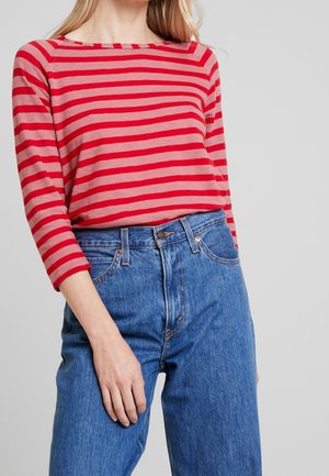 Camicia a righe rossa e beige con scollatura ampia, abbinata a jeans in denim blu a vita alta. Maniche lunghe con polsini arrotolati.