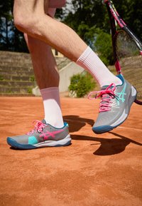 Scarpe da ginnastica grigie con dettagli rosa e blu, parte superiore testurizzata e suola in gomma, abbinate a calzini bianchi fino alla caviglia, su un campo in terra battuta.