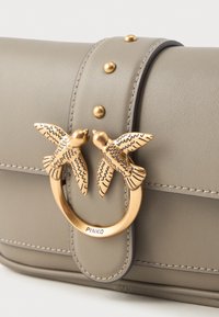 Bolsa de couro taupe com fecho em tom dourado, apresentando dois pássaros detalhados a olhar um para o outro e três studs dourados na alça.