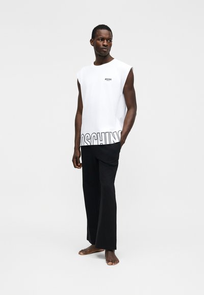 Άνδρας όρθιος ξυπόλυτος, φοράει λευκό αμάνικο τοπ Moschino με μαύρο λογότυπο και χαλαρό μαύρο παντελόνι μπροστά σε απλό ανοιχτό φόντο.