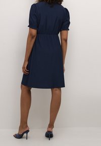 Abito blu navy a maniche corte con vita cintrata, caratterizzato da una texture liscia e una gonna fluida. Abbinato a sandali con tacco blu.