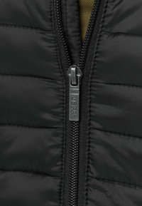 Svart vadderad jacka av tyg med horisontell sömnad; har en frontdragkedja med ett märkt draghandtag och texturerat svart dragkedjeband.