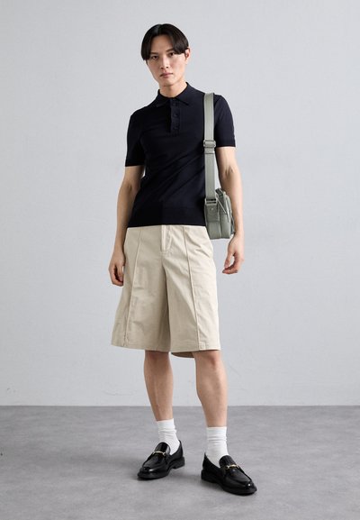 Polo-shirt à manches courtes marine, shorts beige arrivant aux genoux, mocassins noirs et un sac à épaule vert, le tout sur un fond neutre.