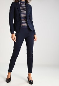 Blazer marine avec une coupe ajustée et un bouton unique, associé à un col roulé à rayures et un pantalon slim, complété par des mocassins noirs brillants.