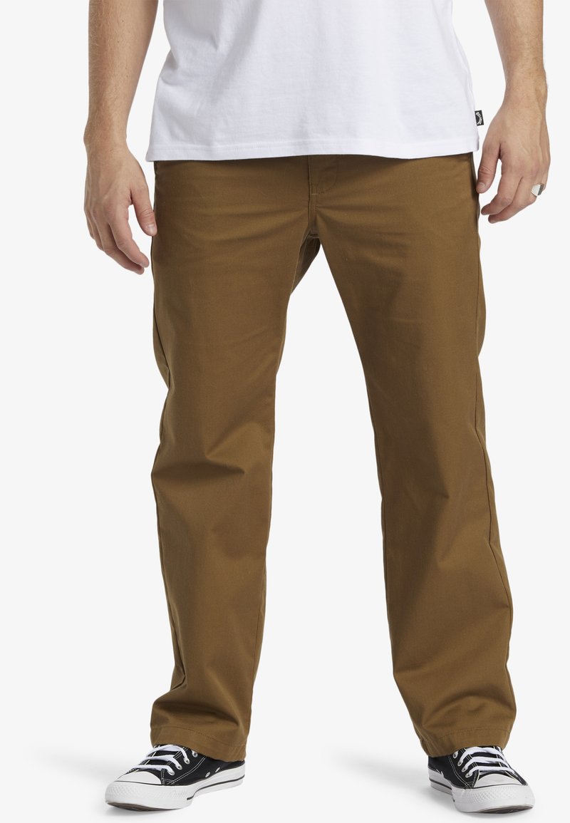 Braune Baumwollhose im geraden Schnitt, mit frontalen Taschen und einer glatten Textur, kombiniert mit schwarz-weißen Turnschuhen.