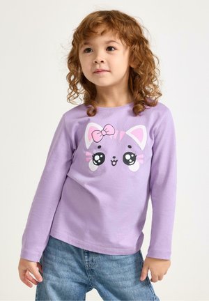 Pitkähihainen violetti puuvillapaita, jossa on kissan kasvokuvio vaaleanpunaisella rusetilla ja yksityiskohtaiset isot silmät, yhdistetty sinisiin farkkuihin.