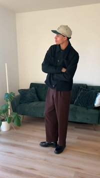 Giacca nera con bottoni, pantaloni marroni plissettati, scarpe nere e berretto grigio. In piedi davanti a un divano verde con cuscini decorativi.