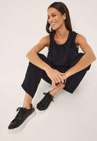 Top senza maniche nero con scollo arricciato, abbinato a pantaloni neri larghi. Le sneakers nere presentano dettagli testurizzati e suole bianche.
