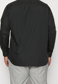 Zwart shirt met lange mouwen en een gladde textuur, voorzien van een kraag, knopen en een iets langere achterkant, gecombineerd met grijze geruite broek.