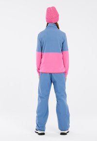 Fleece jas in blauw en roze, met hoge kraag, gecombineerd met blauwe broek. Heeft een zachte textuur en elastische boorden. Draagt een roze gebreide muts.