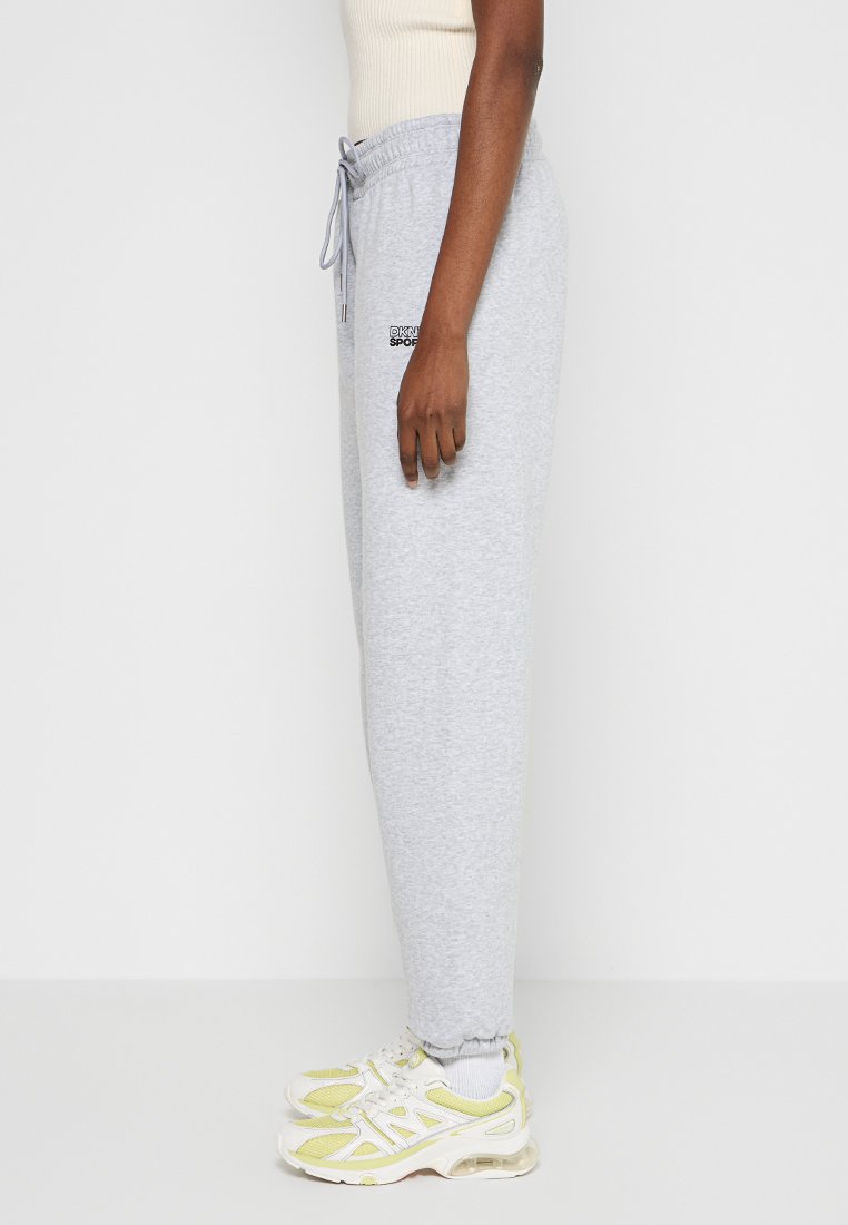 Szare dresy z elastycznym paskiem w talii i sznurkiem, z małym czarnym logo z boku. Noszone z białymi i żółtymi sneakersami.