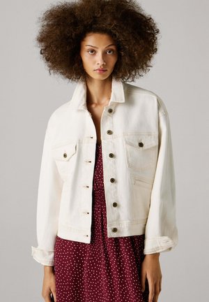Hvit denimjakke med krage, to brystlommer og gullfargede knapper. Brukt over en burgunder kjole med hvite polkaprikker.