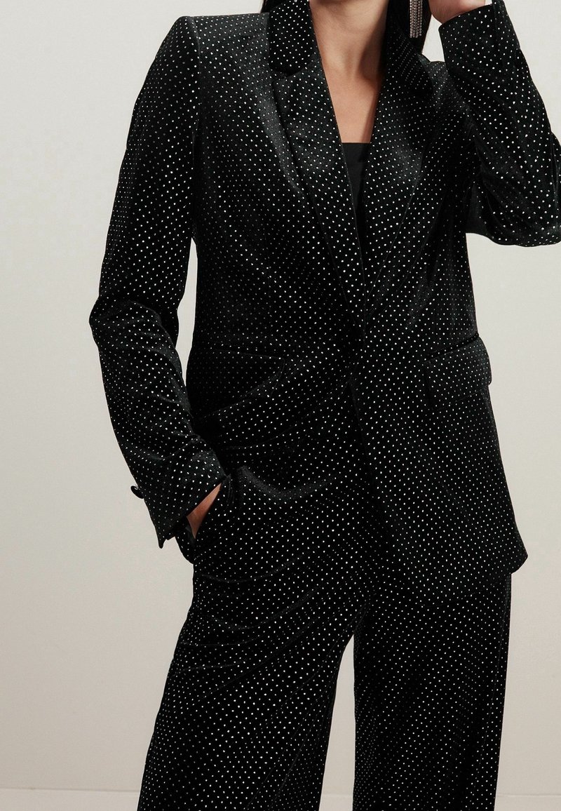 Costume en velours noir à pois blancs. Comprend une veste cintrée avec un col à revers et des poches, assortie à un pantalon coordonné.