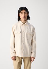 Light beige, textured button-up jacket with two chest pockets and a straight cut, paired with a turtleneck and beige pants.