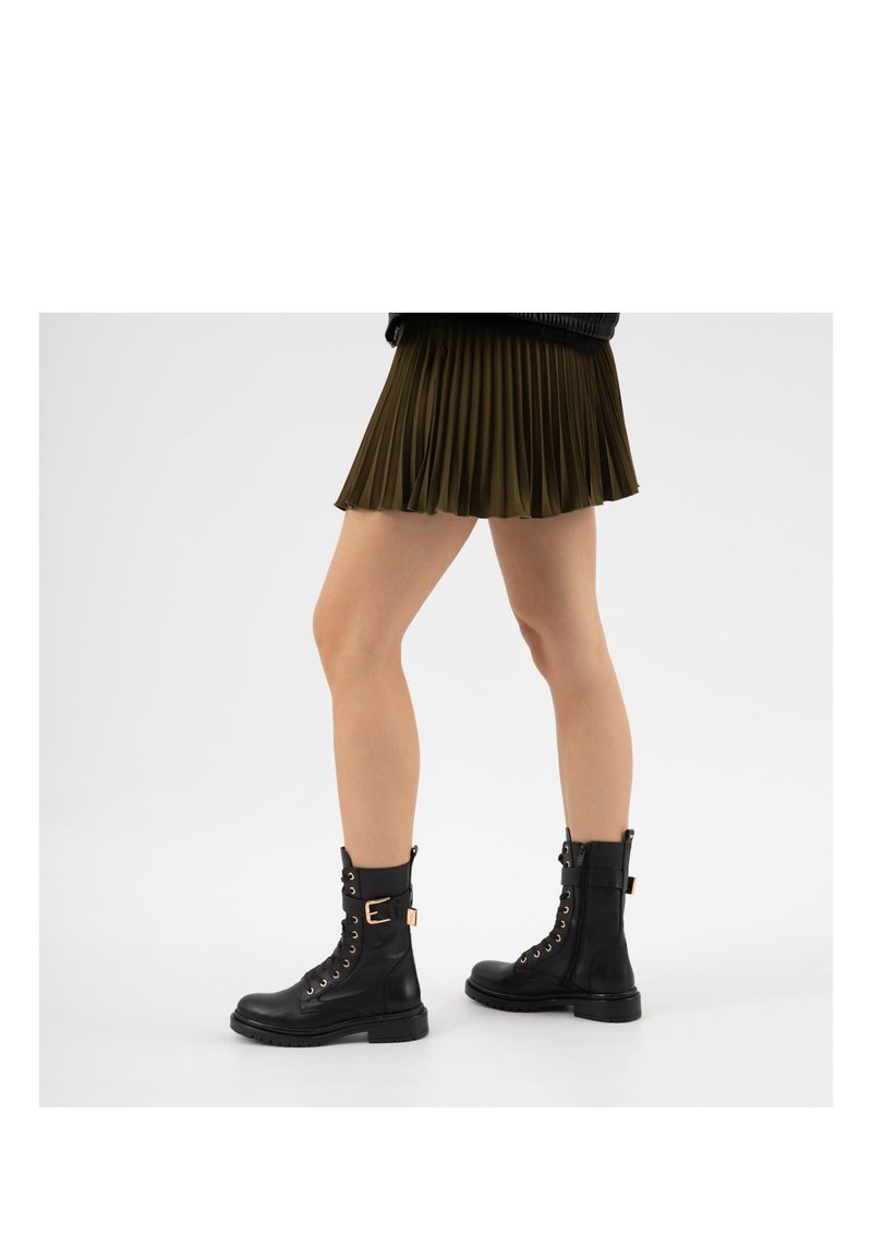 Nohy v olivově zelené plisované sukni a černých šněrovacích vojenských botách s přezkami, stojící na bílém povrchu.