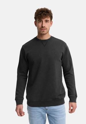 Man met kort krullend haar, gekleed in een effen zwarte longsleeve trui en lichtblauwe spijkerbroek, staand tegen een witte achtergrond.