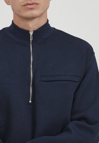 Marinefarbener Reißverschluss-Pullover mit hohem Kragen, ausgestattet mit einem silbernen Reißverschluss und einer Brusttasche. Weicher, strukturierter Stoff, lässige Passform, minimalistisches Design.