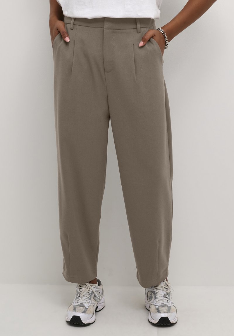 Pantalon taille haute fuselé couleur taupe, porté avec des baskets blanches et un haut blanc rentré, mains posées négligemment dans les poches sur un fond uni.