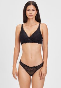 Musta pitsinen bralette, jossa on aaltoilevat reunat ja edessä naru, yhdistettynä vastaavaan mustaan läpinäkyvään stringiin, jossa on kukkakuvioisia pitsikoristeita.