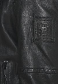 Zwarte leren jas met een ritssluiting, gestructureerd materiaal en een rechthoekige patch met het Strellson-logo op de linkermouw.