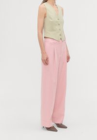 Personne portant un gilet beige sans manches à boutons, un pantalon large taille haute rose et des chaussures pointues gris foncé, debout de profil.