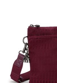 Vinröd crossbody-väska i texturerat tyg, med dragkedjestängning, justerbar rem och ett charmigt hänge fäst vid metalldetaljer.