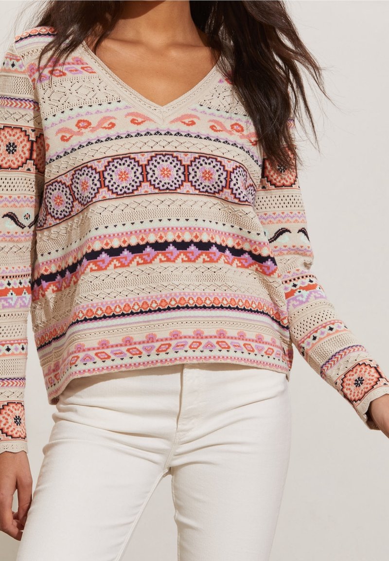 Pull-over en tricot à col en V, présentant des motifs et textures multicolores, principalement en beige, rose et noir, avec des détails complexes.