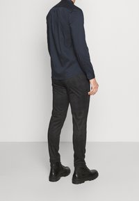 Chemise bleu marine à manches longues et pantalon texturé foncé, associés à des bottes noires à lacets, vus de dos sur un fond uni.