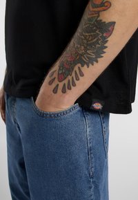 Black short-sleeve t-shirt, relaxed fit, paired with blue denim jeans. Notable tattoo design on the forearm, with a visible brand label on the shirt.