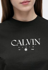 Tânără purtând un tricou negru Calvin Klein cu textul logo alb și o grafică mică florală la piept, pe fundal alb.