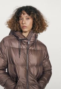 Kvinde med krøllet hår iført en brun dynejakke med høj krave og hænder i lommerne, der ser lige frem.